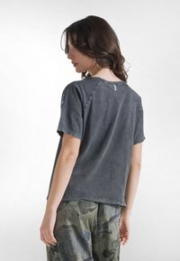 Kvinna med axellångt hår som bär en svartfärgad kortärmad tröja och gröna byxor med blommönster, vänd bort från kameran med huvudet vänt åt vänster.