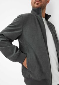Mann trägt eine dunkelgraue Reißverschlussjacke mit gerippten Bündchen und Saum über einem weißen Hemd, die Hand in der Jackentasche.