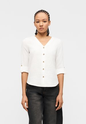 Femme aux cheveux tressés portant une chemise blanche à col en V boutonnée avec les manches retroussées et un jean noir délavé, debout face à l'avant.