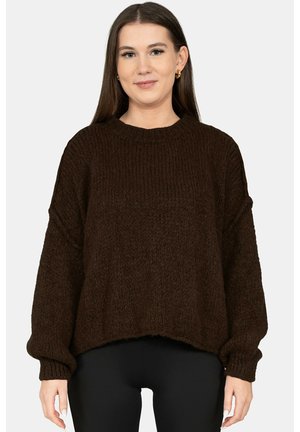 Junge Frau mit langem braunem Haar, die goldene Creolen, einen locker sitzenden dunkelbraunen Strickpullover und schwarze Hosen trägt, vor einem einfarbigen Hintergrund steht.