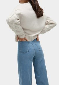Un pull surdimensionné beige clair avec des manches en tricot à torsades, porté avec un jean en denim bleu taille haute avec des poches arrière, texture lisse, coupe décontractée.