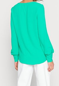 Grüne Bluse mit langen Ärmeln aus leichtem Stoff, mit Rundhalsausschnitt und geknöpften Manschetten, kombiniert mit weißen Hosen.