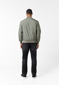 Giacca bomber verde oliva con colletto e polsini a costine, vita elastica, texture liscia, indossata con jeans scuri e sneakers nere.