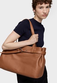 Brązowa skórzana torba typu tote z bocznym ściągaczem, podwójnymi paskami na ramię i subtelnym logo. Gładka faktura, zorganizowany kształt i minimalistyczne detale.