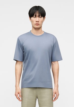 Jeune homme aux cheveux noirs, debout de face, portant un t-shirt uni bleu clair à manches courtes et un pantalon beige sur fond blanc.