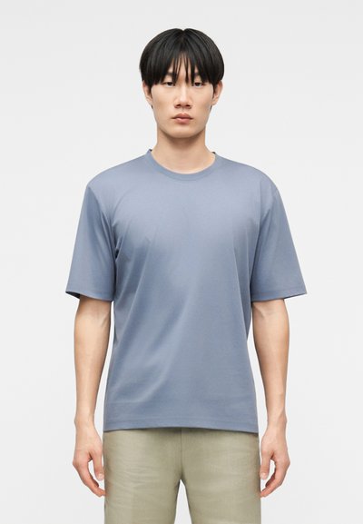 Young man with black hair stands facing forward wearing a plain light blue short-sleeve t-shirt and beige pants against white background.