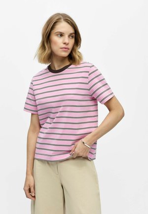 Kvinde med kort blondt hår iført en lyserød stribet T-shirt og beige bukser, stående med den ene hånd i lommen mod en hvid baggrund.