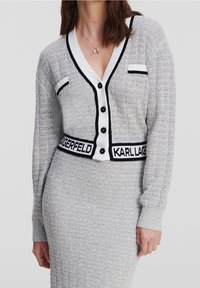 Szary dzianinowy kardigan z dekoltem w kształcie litery V, białymi i czarnymi akcentami oraz charakterystycznym paskiem na talię, z czarnymi guzikami i teksturowaną tkaniną.