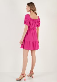 Roze korte jurk met pofmouwen, elastische taille en ronde zoom. De stof lijkt textuur te hebben. Sleehakken met gekleurde accenten.
