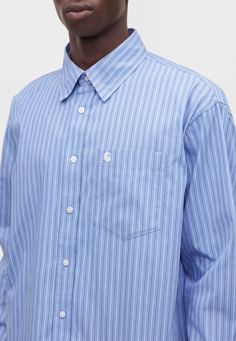 Homme portant une chemise bleu clair à manches longues boutonnée avec des rayures verticales blanches et bleu marine et une poche poitrine avec détail du logo.