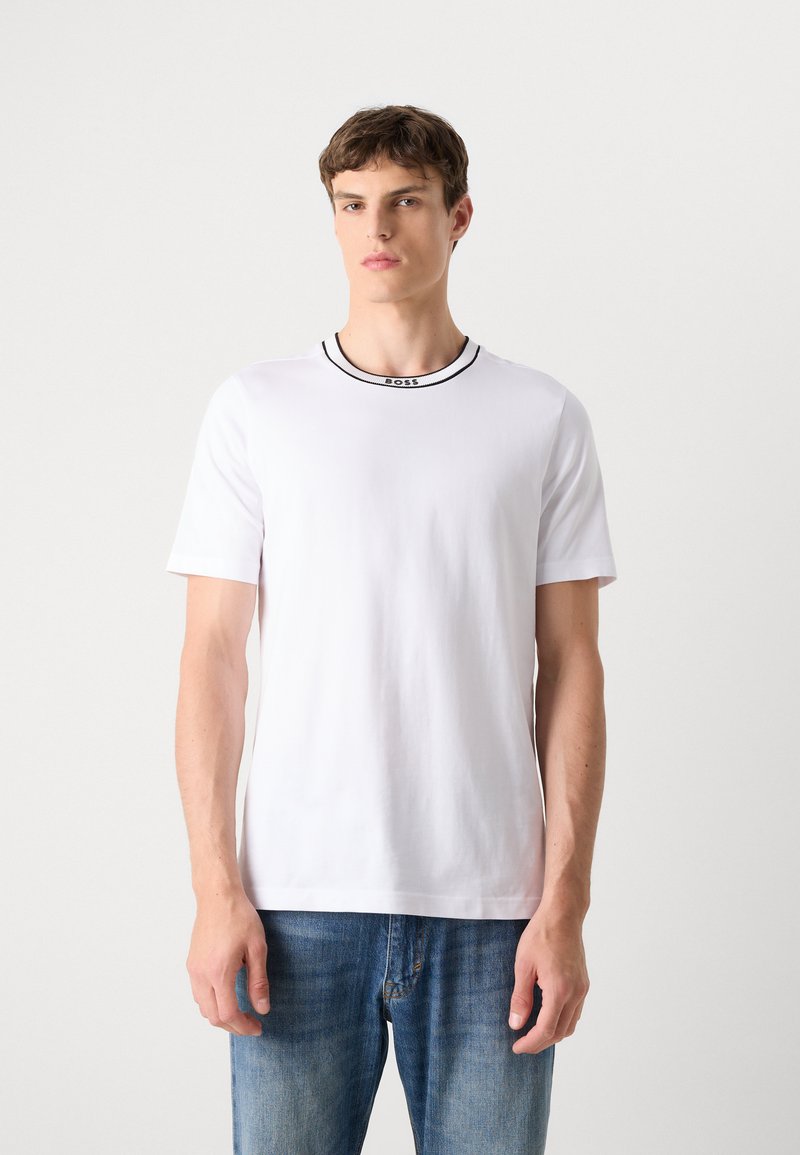 Weißes T-Shirt mit kurzen Ärmeln aus glattem Baumwollstoff, mit einem Kragen in Schwarz und einem Logodetail am Hals. Getragen mit blauen Jeans.