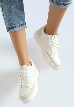 Feet wearing white sneakers with beige accents and rolled-up blue jeans against a plain light background.