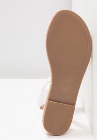 Semelle d'une sandale plate beige avec une semelle en caoutchouc texturée et un bord tissé, positionnée à la verticale contre une surface blanche.