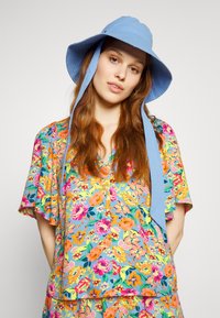 Chapéu de palha de aba larga em azul claro, com fitas para o pescoço, combinado com uma blusa de manga curta vibrante, com padrão floral nas cores azul, rosa e amarelo.