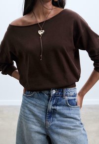 Pull marron à épaules dénudées avec manches longues, associé à un jean bleu clair taille haute. Collier notable avec pendentif en forme de cœur sur un cordon en cuir.