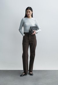 Svetlosivý rebrovaný top s dlhým rukávom v kombinácii s hnedými nohavicami s vysokým pásom, držiac malú čiernu kabelku s cvokmi. Čierne balerínky.