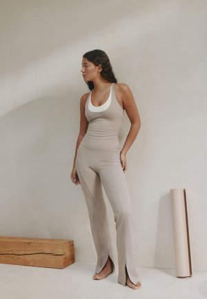 Frau in beigem, tailliertem Tanktop und hoch taillierter Hose mit Schlitz am Saum steht barfuß auf hellem Boden in der Nähe einer Yogamatte und eines Holzblocks.