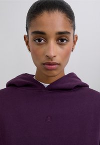 Fioletowa bluza z kapturem o fakturowanej tkaninie, z małym haftowanym logo czaszki. Kaptur jest luźny, a bluza jest noszona na białej koszuli.