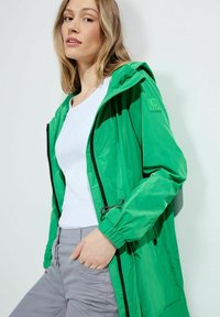 Grön lättviktsjacka med huva, dragkedjestängning och elastiska muddar. I kombination med en vit topp och grå byxor. Slät textur. C-logotypdetalj.