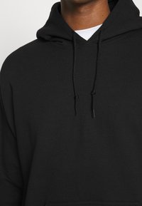 Sort bomulds hoodie med snoretræk i hætten, en lomme foran og en blød tekstur. Designet er enkelt og afdæmpet.