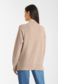 Beige gebreide trui met een hoge ribbelnek en een relaxte pasvorm, met lange mouwen en ribbelmanchetten en zoom. Gecombineerd met blauwe jeans.