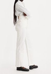 Jeans bianchi a vita alta con gamba dritta, caratterizzati da orlo grezzo e accessori in rame, abbinati a mocassini neri e una camicia bianca oversize.