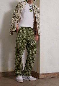 Pantalon vert à motif avec texte blanc, associé à une veste imprimée florale, une chemise blanche et des baskets blanches, devant un mur neutre.
