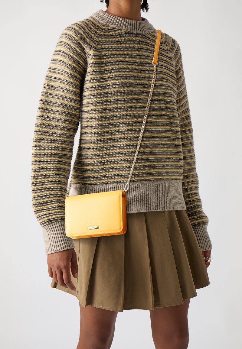 Personne portant un pull en maille rayé et une jupe plissée verte, portant un petit sac bandoulière jaune avec une chaîne.