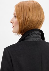 Frau mit kurzem roten Haar, die einen schwarzen Mantel mit hochgestelltem Kragen trägt, auf dem das Logo "S.OLIVER" auf weißem Hintergrund zu sehen ist.