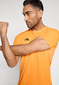 Oranges Sport-T-Shirt mit kurzen Ärmeln, das auf der Brust ein schwarzes Drei-Streifen-Logo zeigt und eine glatte, leichte Stofftextur aufweist.