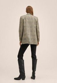 Blazer à carreaux gris avec une coupe ajustée, associé à un pantalon noir slim et des bottes noires montant jusqu'au genou agrémentées d'un motif floral.