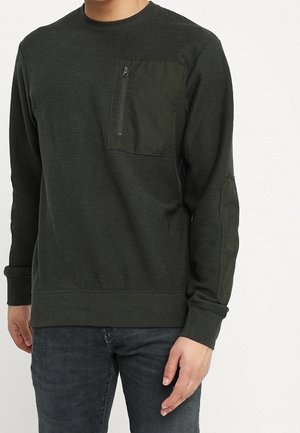 Homme portant une chemise à manches longues texturée vert foncé avec poche poitrine zippée et coudières, associée à un jean bleu foncé.