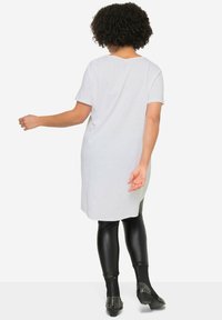 Witte T-shirt tot de knie met korte mouwen en een ronde halslijn, gecombineerd met zwarte, glanzende leggings en zwart, gespiked enkellaarzen.