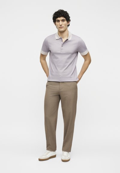 Νέος άνδρας στέκεται με τα χέρια πίσω από την πλάτη, φοράει ανοιχτό μωβ πόλο μπλούζα, μπεζ παντελόνι και λευκά αθλητικά παπούτσια σε απλό λευκό φόντο.