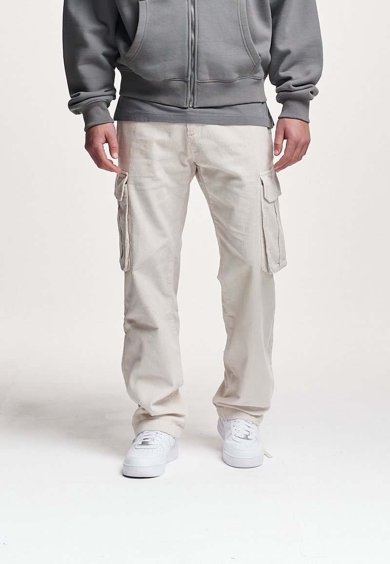 Homme portant un pantalon cargo beige, des baskets blanches et un sweat à capuche gris zippé, debout sur un fond blanc.