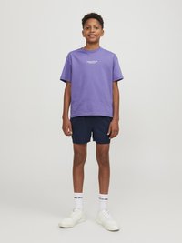 Paarse katoenen t-shirt met korte mouwen, voorzien van een witte tekstafbeelding. Geroid met marineblauwe shorts en witte sneakers, met zichtbare branding op de sokken.