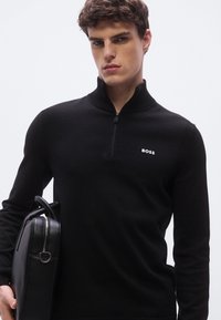 Svart genser med glidelås, ribbestrikkede detaljer og en logo på brystet, kombinert med en svart lærmappe. Jevn tekstur og tettsittende design.