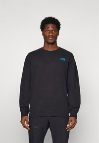 Man in een zwarte crewneck sweatshirt met een klein felblauw The North Face-logo op de linkerborst, staand tegen een effen achtergrond.