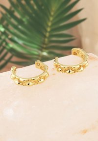 Guldörhängen i form av ringar har en scalloped design med rundade accenter. De vilar på en ljus stenytan med en suddig grön bakgrund.