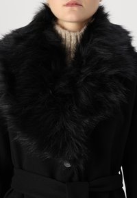 Sort frakke med en bred krave af kunstpels. Kraven har en blød, fluffy tekstur. Frakken har et bælte og en knaplukning.
