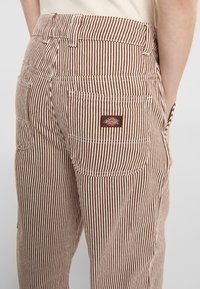 Braun-weiße gestreifte Arbeitshose mit Gesäßtaschen und einem kleinen Dickies-Logopatch. Hergestellt aus strapazierfähigem Stoff mit strukturierter Oberfläche.