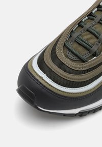 Olivově zelené a černé tenisky s síťovinovými panely, vrstveným designem a kontrastními bílými akcenty; obsahují texturované povrchy a gumovou podrážku.