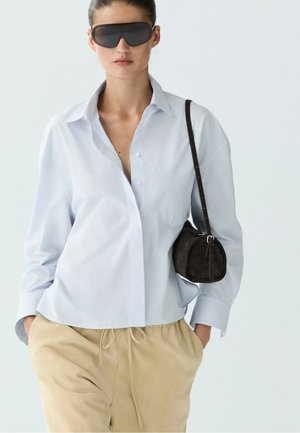 Donna con camicia oversize azzurro chiaro con bottoni, pantaloni beige con coulisse, grandi occhiali da sole neri e piccola borsa a tracolla marrone scuro.