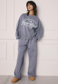 Grå sweatshirt med hvid tekst, matchende grå løstsiddende bukser og tan slip-on fodtøj. Blødt stof, afslappet design, minimalistisk stil.