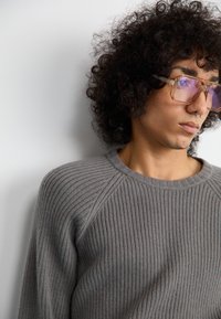 Siv pulover s rebrasto teksturo, kroja s krožnim izrezom in raglan rokavi; model nosi oversized prozorne očala z svetlo vijoličnimi lečami.