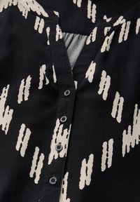 Blusa nera con scollatura a V, motivo astratto color beige chiaro e sei bottoni neri sul davanti con una texture liscia e lucida.