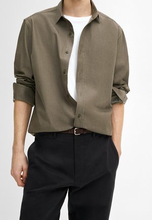 Man met een opengeknopte olijfgroene overhemd over een wit T-shirt, zwarte broek, bruine riem, mouwen opgerold, staand met één hand in de zak.