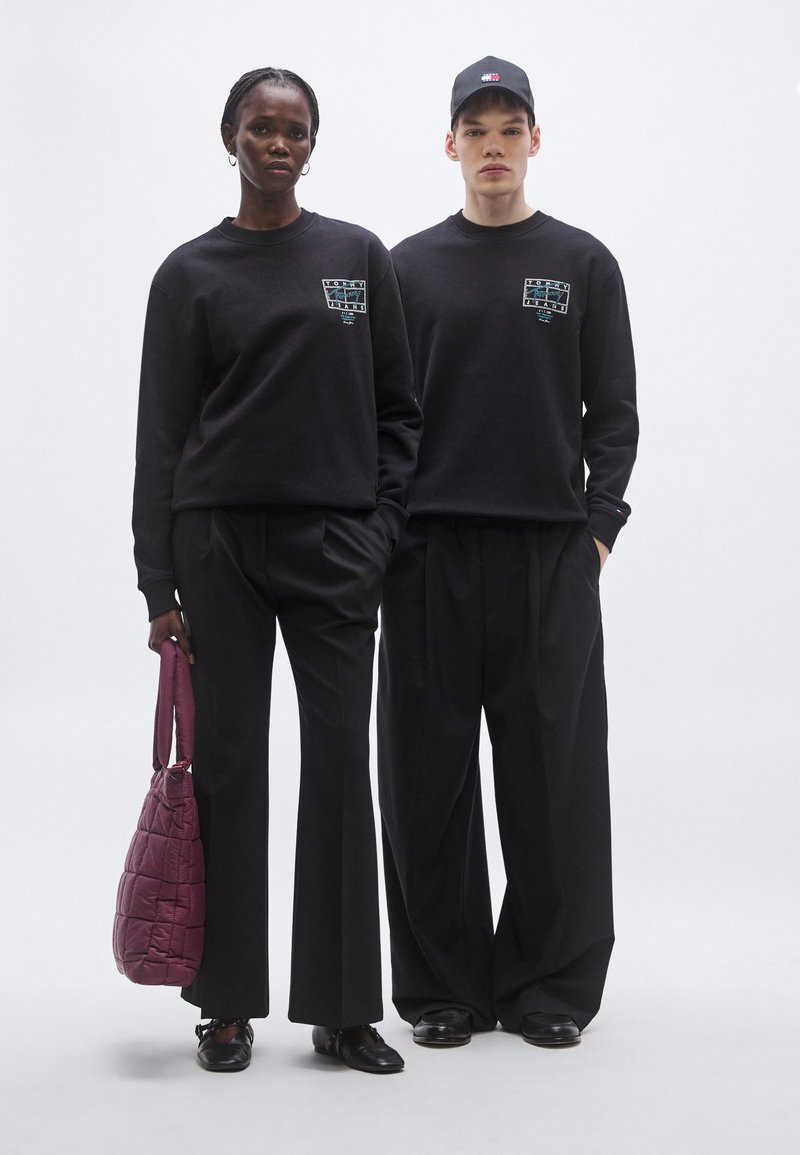 Two individuals wear matching black sweatshirts with graphic details. The woman holds a burgundy quilted handbag, paired with black trousers and shoes.