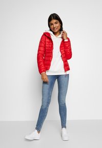 Mujer sonriendo, vistiendo una chaqueta acolchada roja sobre un sudadera blanca, jeans ajustados de color azul claro y zapatillas blancas, de pie contra un fondo blanco liso.