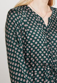 Groene gebloemde blouse met kleine bloemenpatronen, lange mouwen, een V-hals en decoratieve knopen aan de voorkant. Zachte stof.